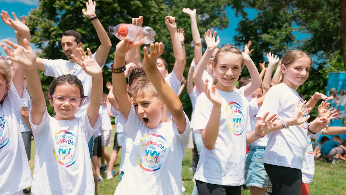 Group of cheerful children raising hands in a park during a sunny day, wearing t-shirts with event logos.